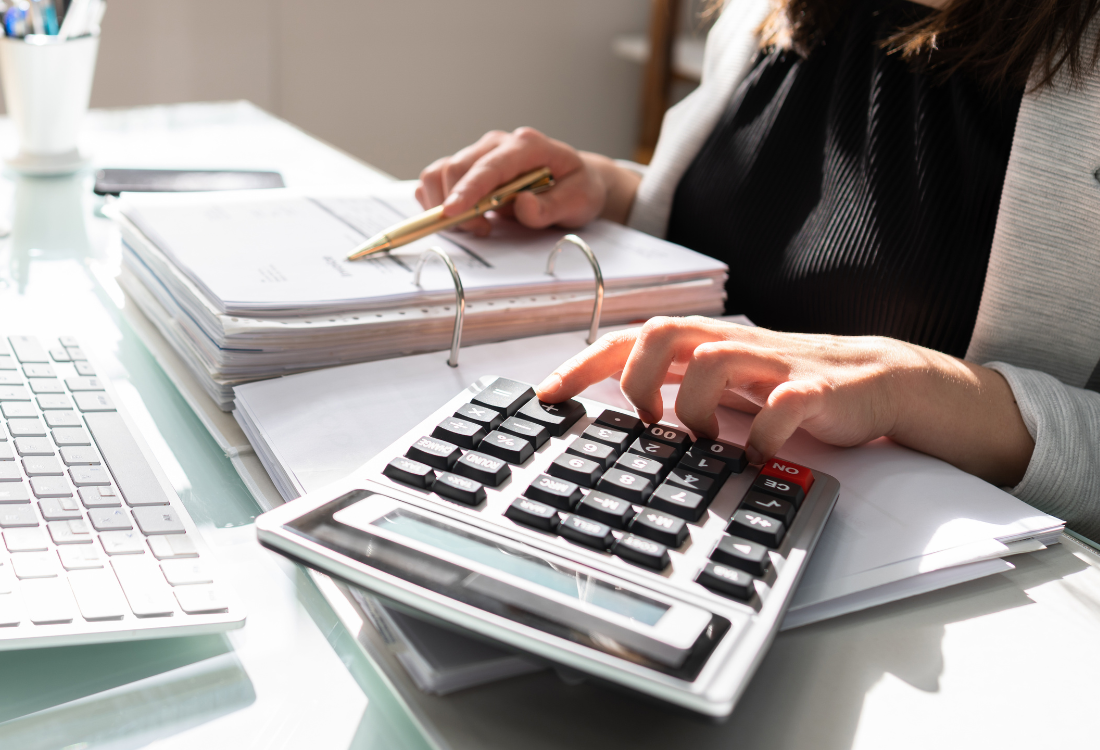 A female accountant, sat at her desk using a calculator and looing at her book whilst using accounting software for small businesses